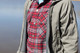 Man wearing a flannel shirt, green jacket on beach, next to water.